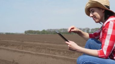 Kadın çiftçi, tarlada özel toprak sıralarının arasında oturuyor. Test ediyor, tablet kullanıyor, ekicinin patates ekme kalitesini ölçüyor. Modern tarımsal olarak patates yetiştirme. Bahar güneşli gün.