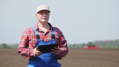 Çiftçi, tarım uzmanı, tarlada duruyor, testler, tablet kullanımı, modern tarımın kalitesi. Bahar güneşli bir gün. Çalışan traktörün arkaplanı, tarlada yetiştirici