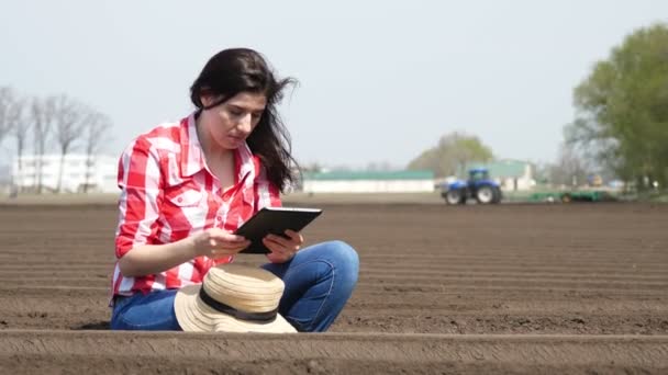 Agricultora, agrónoma se sienta entre hileras especiales de suelo en el campo. Ella prueba, usando tableta, la calidad de la plantación de patata por cultivador. Moderno cultivo de patata agrícola. Primavera día soleado . — Vídeos de Stock