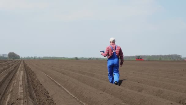 Boer, agronomist loopt op het veld met speciale bodem rijen. Hij test, met behulp van tabletten, de kwaliteit van aardappelplanten door cultivator. Moderne teelt. zonnige lentedag. — Stockvideo