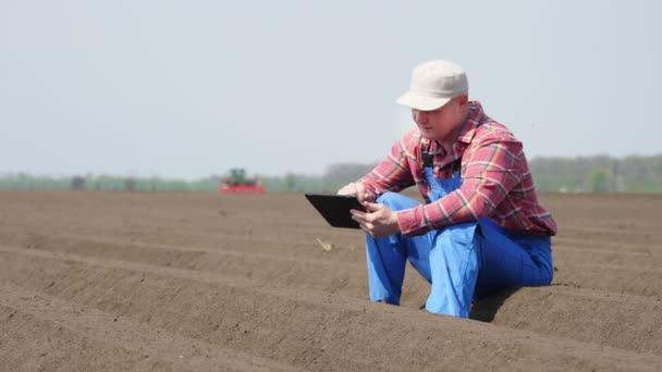 Фермер, агроном сидить між спеціальними рядами ґрунтів на полі. він аналізує, використовуючи таблетки, якість посадки картоплі культиватором. Сучасне сільськогосподарське вирощування картоплі. весняний сонячний день . — стокове відео