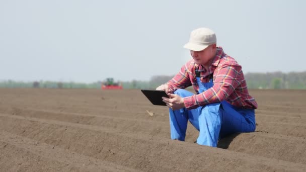 Agricultor, agrónomo se sienta entre hileras especiales de suelo en el campo. prueba, usando tableta, la calidad de la plantación de patata por cultivador. Moderno cultivo de patata agrícola. Primavera día soleado . — Vídeos de Stock