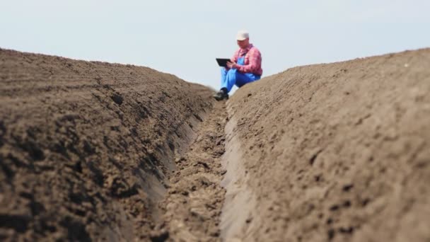 Farmer, agronomist sits between special soil rows on field. he tests, using tablet, quality of potatoe planting by cultivator. Modern Agriculturally potato cultivation. spring sunny day. — Stock Video