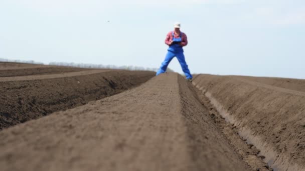 Jordbrukaren står på fältet, mellan speciella jordrader. Han testar potatisplanteringens kvalitet med hjälp av surfplatta. Modern jord- och skogsbruk. vår solig dag. — Stockvideo
