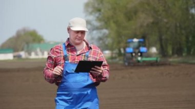 Çiftçi, tarım uzmanı, tarlada duruyor, testler, tablet kullanımı, modern tarımın kalitesi. Bahar güneşli bir gün. Çalışan traktörün arkaplanı, tarlada yetiştirici