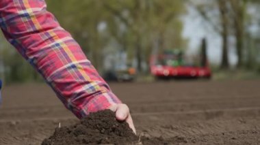 Yakın plan, çiftçi, tarım uzmanı tarladaki toprağın kalitesini kontrol ediyor. Çalışan traktör ve ekicinin arka planı. Modern tarımsal olarak patates yetiştirme. Bahar güneşli gün.