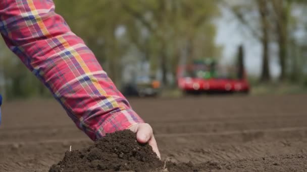 Yakın plan, çiftçi, tarım uzmanı tarladaki toprağın kalitesini kontrol ediyor. Çalışan traktör ve ekicinin arka planı. Modern tarımsal olarak patates yetiştirme. Bahar güneşli gün. — Stok video