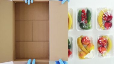 Üst Manzara, kapat. Koruyucu eldivenlerdeki gönüllüler bakkal paketleri, meyve ve sebzeleri yiyecek kutusuna koyarlar. Hayırseverlik. Coronavirus sırasında paket servis, kapı yemek servisi. İnternetten alışveriş.