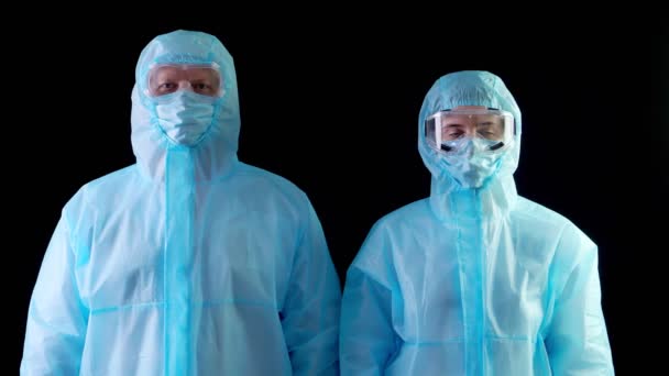 Medisch personeel, dokter en verpleegkundige, in speciale beschermende uniformen, maskers, brillen, kijkend naar de camera. Coronavirusepidemie. op zwarte achtergrond. Bescherming van het coronavirus — Stockvideo