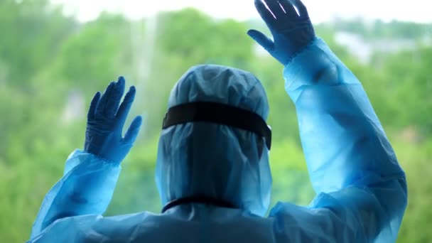 Özel koruyucu giysili bir sağlık çalışanının arka görüntüsü. Yorgun ve bitkin doktor pencereye yaslandı. Koronavirüs salgını, covid-19 salgını. koruma — Stok video