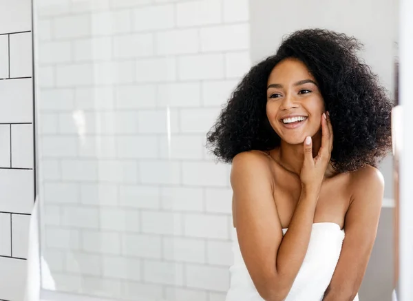 Smiling woman observing her face in mirror, standing in bathroom in front of mirror