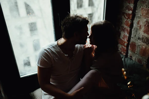 Feliz pareja de amantes en pijama se sientan en el alféizar de la ventana. Ambiente navideño . —  Fotos de Stock