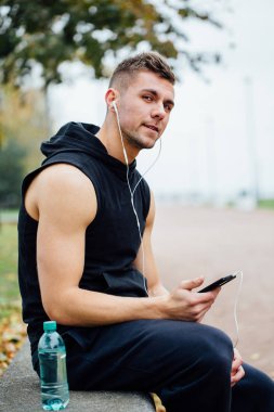 Fitness spor taş bir bankta oturan müzik dinlemek giyen adam. Konuşmak zor egzersiz yaptıktan sonra rahatla.