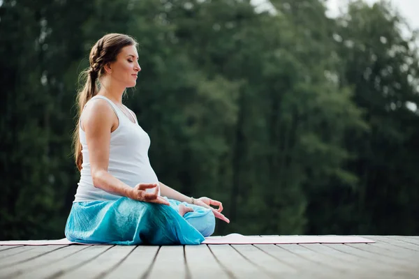 Genç kadın lotus pozisyonunda Nehri yanındaki ormanda yoga pratiği yapıyor. ahşap iskele paspaslar üzerine oturan. — Stok fotoğraf