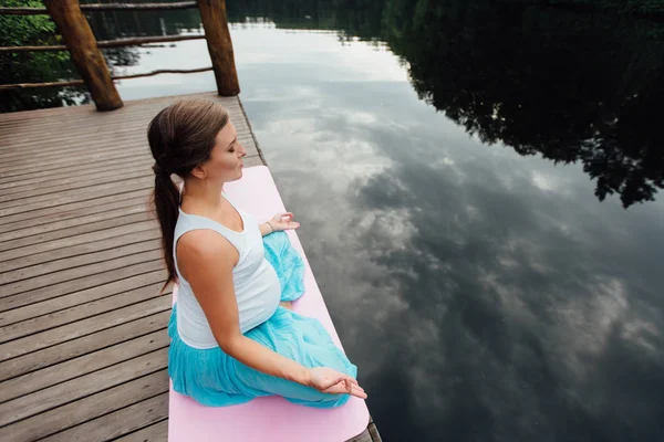 Young woman in the lotus position is practicing yoga in the forest next to the river. sitting on mats  the wooden pier. — Stock Photo, Image