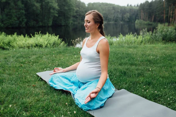 Yoga embarazada en la posición de loto en el fondo del bosque. en el parque la alfombra de hierba, al aire libre, mujer de la salud . — Foto de Stock