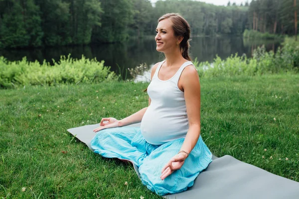 Беременная йога в позе лотоса на фоне леса. в парке коврик для травы, открытый, женщина здоровья . — стоковое фото