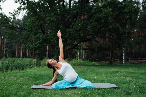 Schwangerschaftliches Yoga in der Lotusposition vor dem Hintergrund des Waldes. im Park die Rasenmatte, im Freien, Gesundheit Frau. — Stockfoto