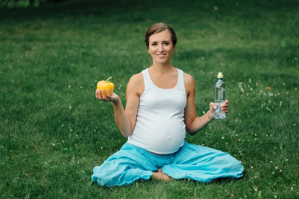 Mujer embarazada de yoga con una botella de agua y pimiento, en la posición de loto. Parque, hierba. al aire libre, bosque . — Foto de Stock