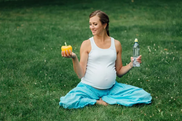 Mujer embarazada de yoga con una botella de agua y pimiento, en la posición de loto. Parque, hierba. al aire libre, bosque . — Foto de Stock