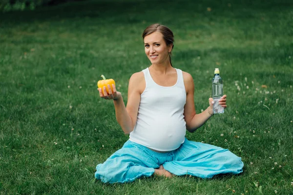 Mujer embarazada de yoga con una botella de agua y pimiento, en la posición de loto. Parque, hierba. al aire libre, bosque . — Foto de Stock