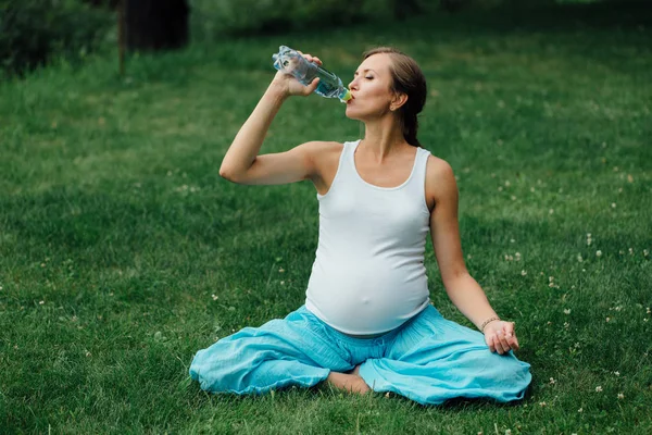Mujer embarazada de yoga bebiendo agua de una botella, en la posición de loto. parque, hierba, .outdoor, bosque . — Foto de Stock