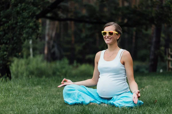 Mujer embarazada de yoga feliz en gafas de sol, en la posición de loto. Parque, hierba. exterior , — Foto de Stock