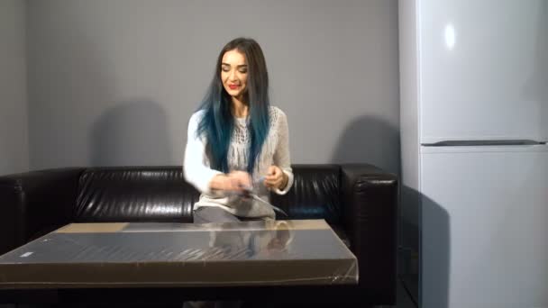 Young cute girl with colored hair sitting on a couch in their new apartment to put together a table. Shoot in kitchen. unpacks the package with scissors — Stock Video