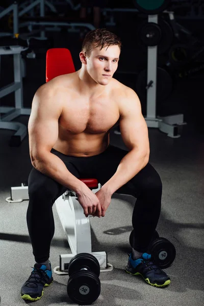 Culturista profesional sentado en el banco, descansando entre ejercicios con pesas en el gimnasio. Grande. — Foto de Stock