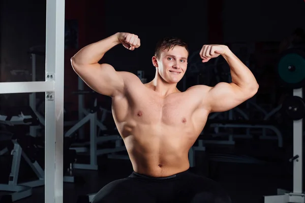 Professional fitness bodybuilder sitting on the bench and demonstrates muscles of your body after a workout, at gym. — Stock Photo, Image