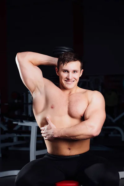 Professionele bodybuilder training met barbell zittend op het bankje op sportschool. Grote zelfverzekerde gespierde man opleiding. — Stockfoto