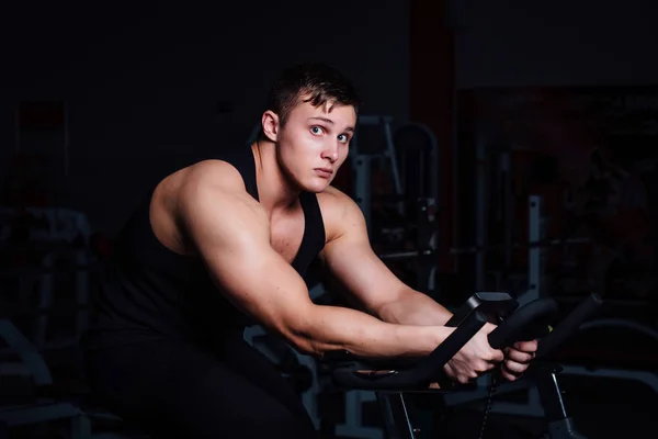 Portrét pohledný muž cvičení na fitness rotopedu tmavé v tělocvičně. — Stock fotografie