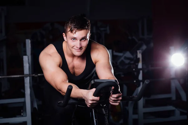 Retrato de un hombre guapo entrenar en fitness la bicicleta de ejercicio oscuro en el gimnasio . — Foto de Stock