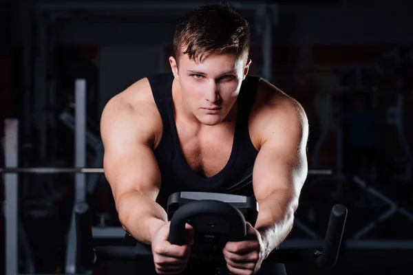 Retrato de un hombre guapo entrenar en fitness la bicicleta de ejercicio oscuro en el gimnasio . — Foto de Stock