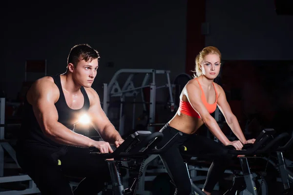 Pareja deportiva haciendo ejercicio en el gimnasio la bicicleta estática en un gimnasio oscuro . — Foto de Stock