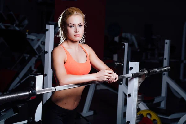Блондинка фитнес-девушка с идеальной формой тела отдыхает после спортивных тренировок в тренажерном зале опираясь на штангу — стоковое фото