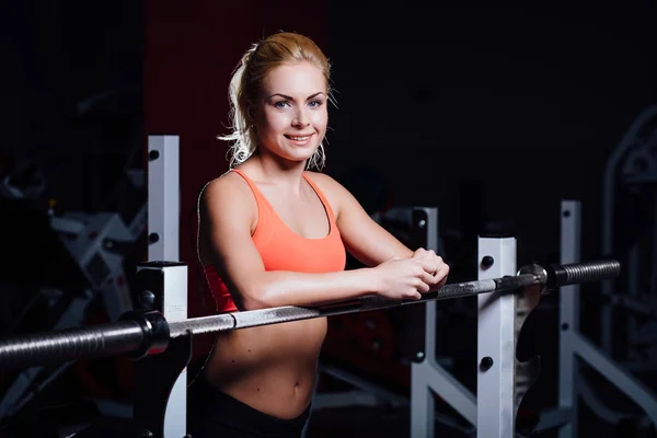 Blonde fitness girl with perfect shape body rests after sport workout in gym leaning on barbell — Stock Photo, Image