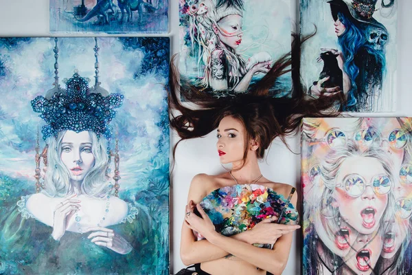 Hermosa artista desnuda femenina rodeada de pinturas lienzo sobre el fondo blanco del suelo. pinceles y paleta . — Foto de Stock