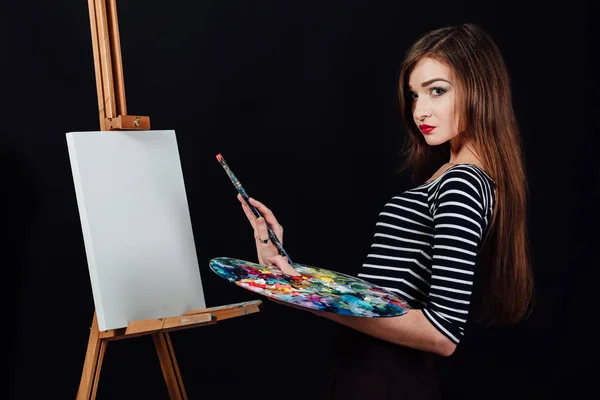 Carino bella ragazza artista pittura un quadro su tela un cavalletto. Spazio per il testo. Studio sfondo nero . — Foto Stock