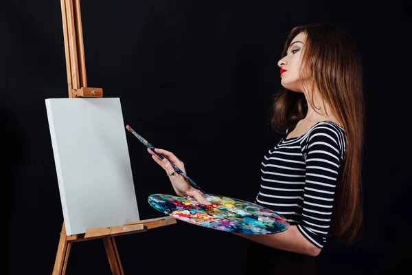 Симпатичная красивая девушка-художник рисует картину на холсте мольберта. Пространство для текста. Чёрный фон . — стоковое фото