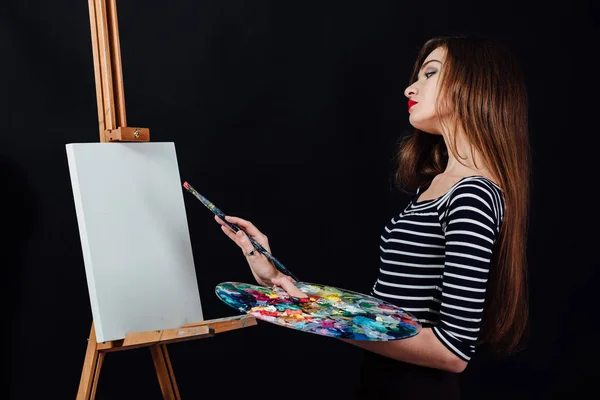 Carino bella ragazza artista pittura un quadro su tela un cavalletto. Spazio per il testo. Studio sfondo nero . — Foto Stock