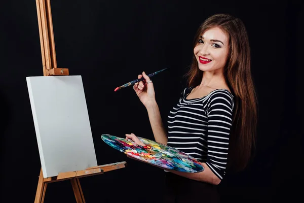 Carino bella ragazza artista pittura un quadro su tela un cavalletto. Spazio per il testo. Studio sfondo nero . — Foto Stock