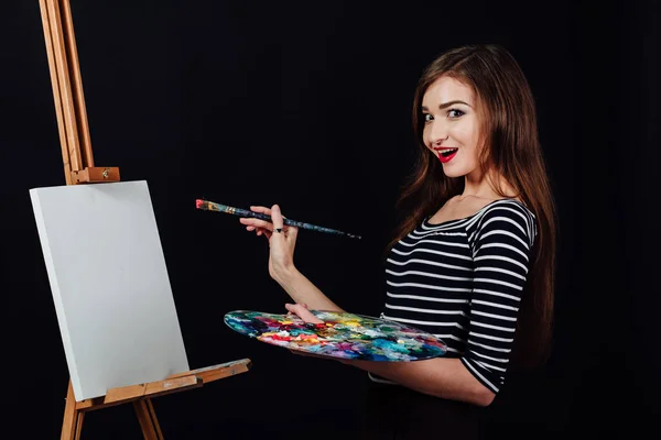Carino bella ragazza artista pittura un quadro su tela un cavalletto. Spazio per il testo. Studio sfondo nero . — Foto Stock