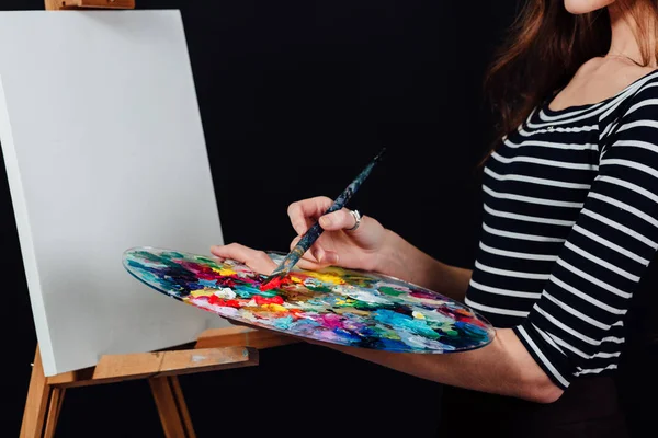 Carino bella ragazza artista pittura un quadro su tela un cavalletto. Spazio per il testo. Studio sfondo nero . — Foto Stock