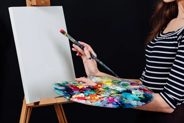 Schattige mooie meisje kunstenaar schilderen van een foto op canvas een ezel. Ruimte voor tekst. Studio zwarte achtergrond. — Stockfoto