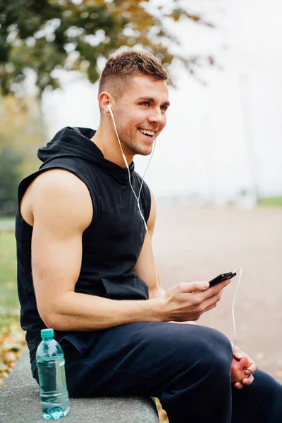 Fitness man wearing sportswear listening to music sitting on a stone bench. Relax after  hard workout, talking.