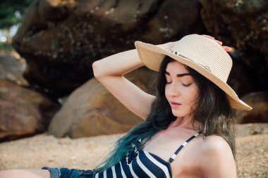 Uzun saçlı kız bir hipster şapka ve çizgili mayo relax tropik Sahilde taş arka plan.