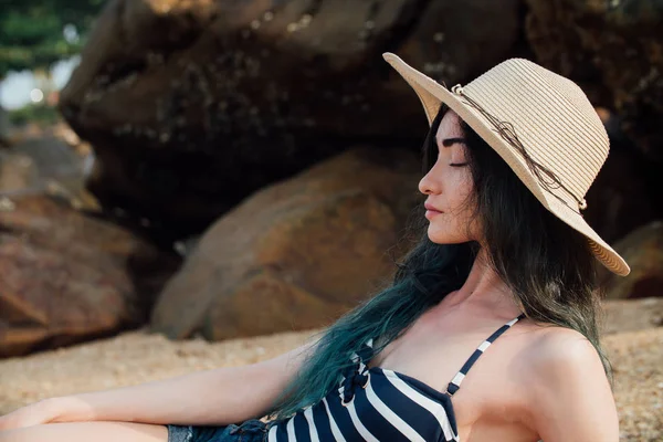 Menina de cabelos compridos Em um chapéu hipster e maiô listrado relaxar na praia tropical, fundo de pedras . — Fotografia de Stock