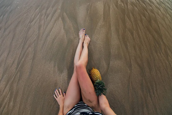Όμορφο μακρύ γυναικεία πόδια στην παραλία, στο χέρι του ένα ανανά, κάτοψη. — Φωτογραφία Αρχείου