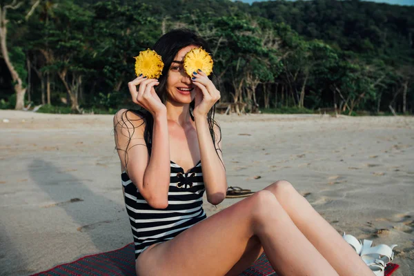 Beautiful young woman tourist In a striped swimsuit holding two ripe pineapple against her eyes. — Stock Photo, Image